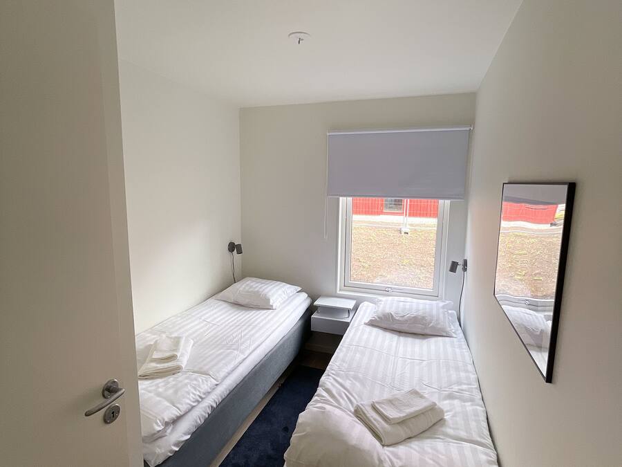 A small, modern room with two single beds, a small table, a window with a view, and a ceiling-mounted smoke detector.