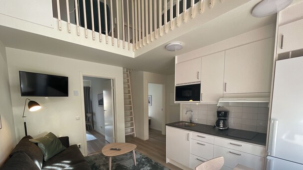 Una sala de estar moderna con un sofá, una mesa de centro y un televisor. Hay una zona de cocina con un microondas, una cafetera y armarios. Una escalera está apoyada contra la pared, y una puerta da a otra habitación.