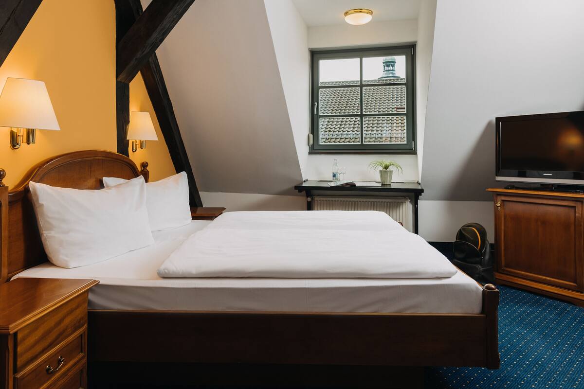 A neatly made bed in a hotel room with a wooden headboard, a nightstand, a lamp, a television, and a window with a view of a building.