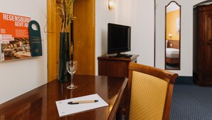 Une chambre d’hôtel avec une table à manger en bois, un vase rempli de fleurs, un verre à vin, un téléviseur posé sur un meuble et un miroir accroché au mur.