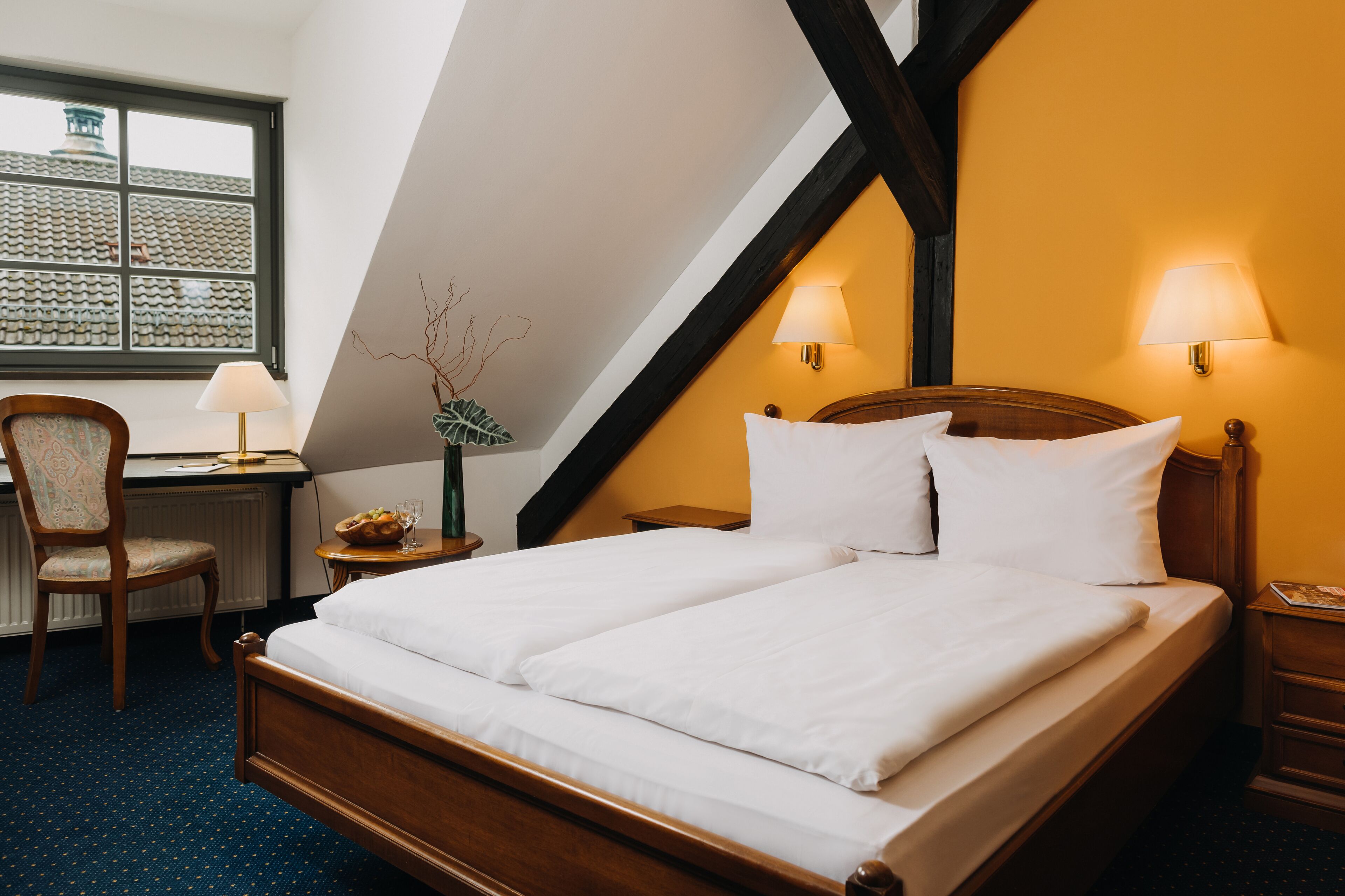 A double bed with white linens, a wooden headboard, two bedside lamps, a chair, a small table with a lamp, and a view of the outside through a window with a tiled roof.