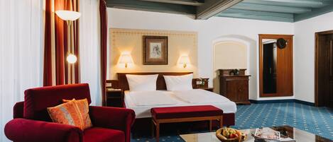 Una habitación de hotel con una cama, un sillón rojo, un banco de madera, una mesa de centro de vidrio con frutas y una revista, y una alfombra azul estampada.