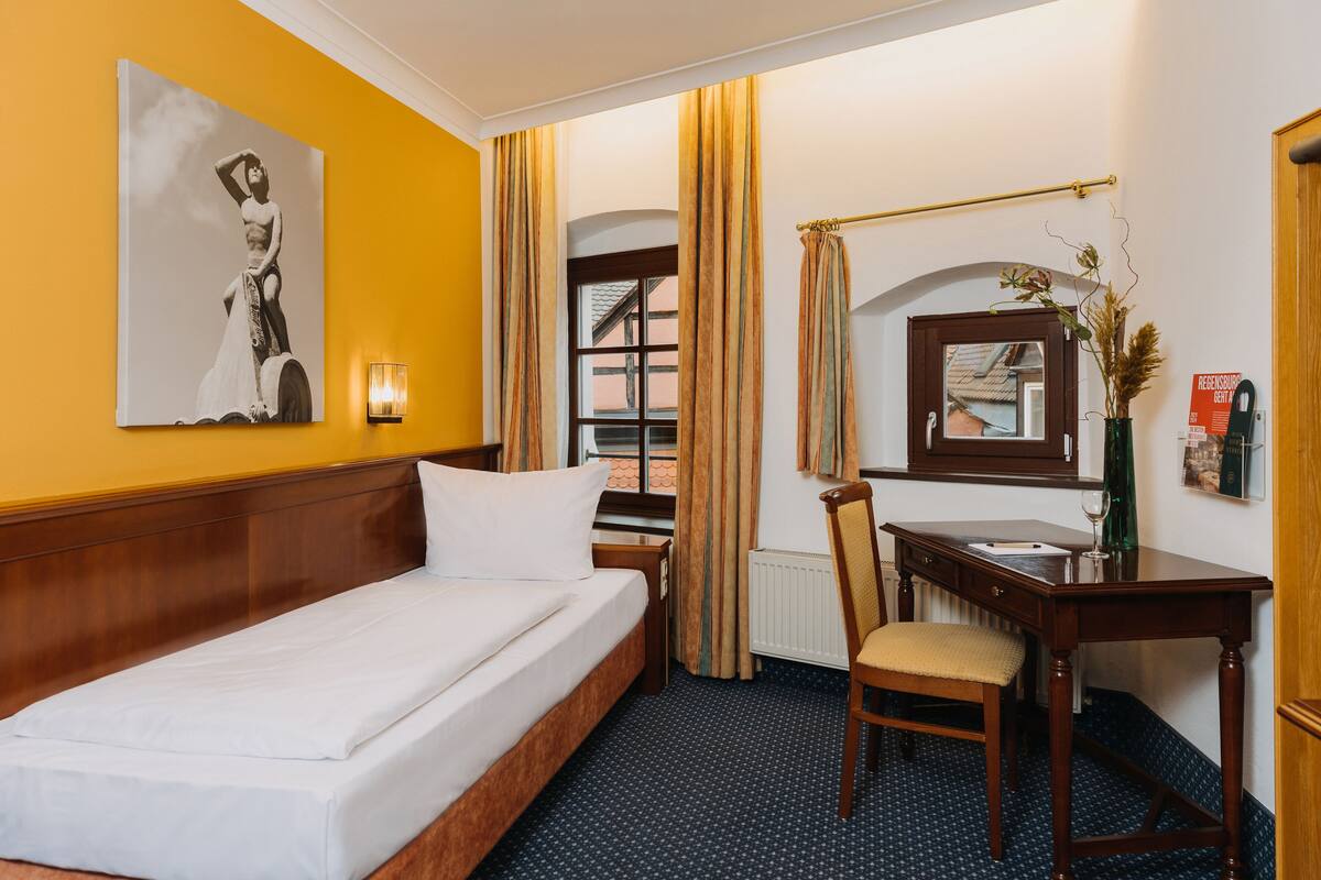 A hotel room with a bed, a desk, a chair, a vase with dried plants, a framed picture on the wall, and a window with curtains.