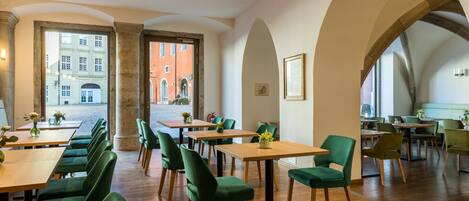 A restaurant with green upholstered chairs, wooden tables, and a view of a red building through an arched doorway.