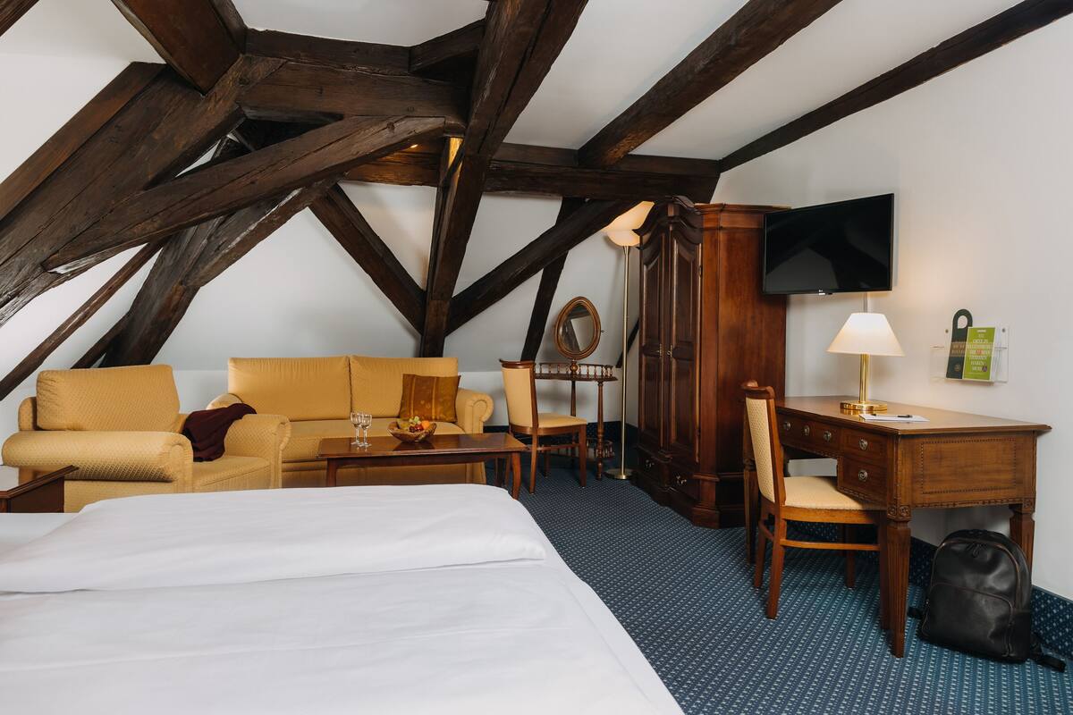 A hotel room with a bed, a sofa, a desk, a chair, a television, and a wooden ceiling structure.