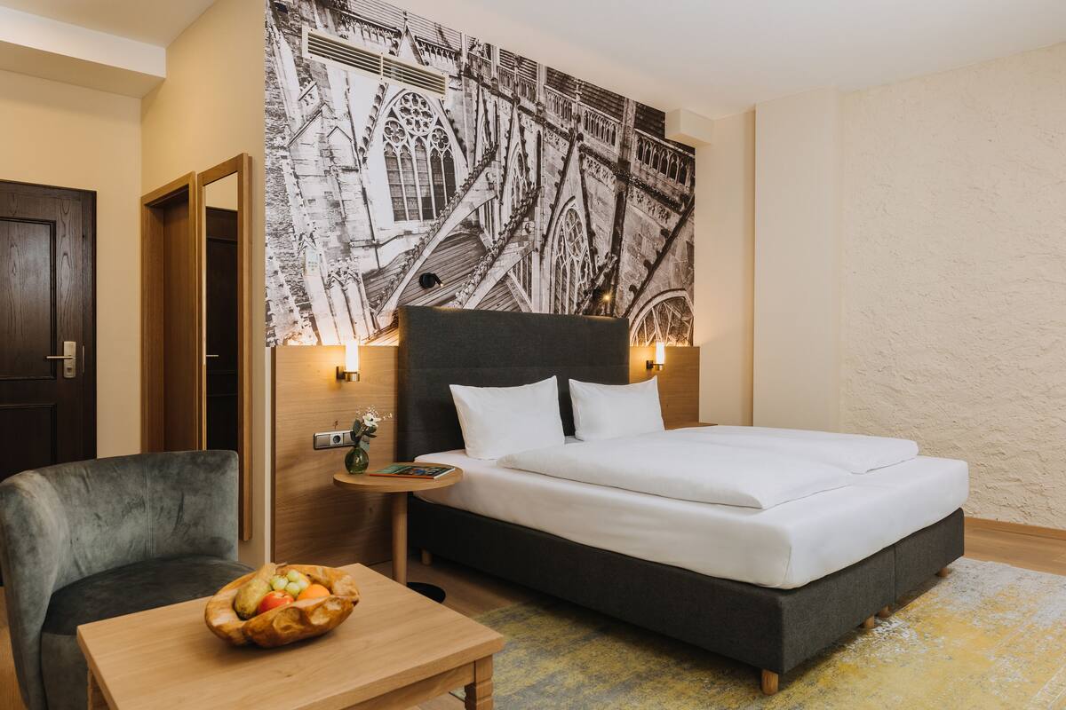 A hotel room with a large bed, a chair, a small table with a fruit bowl, and a wall tapestry.