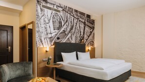Une chambre d’hôtel avec un grand lit, une chaise, une petite table sur laquelle se trouve un panier de fruits, et une tapisserie murale.