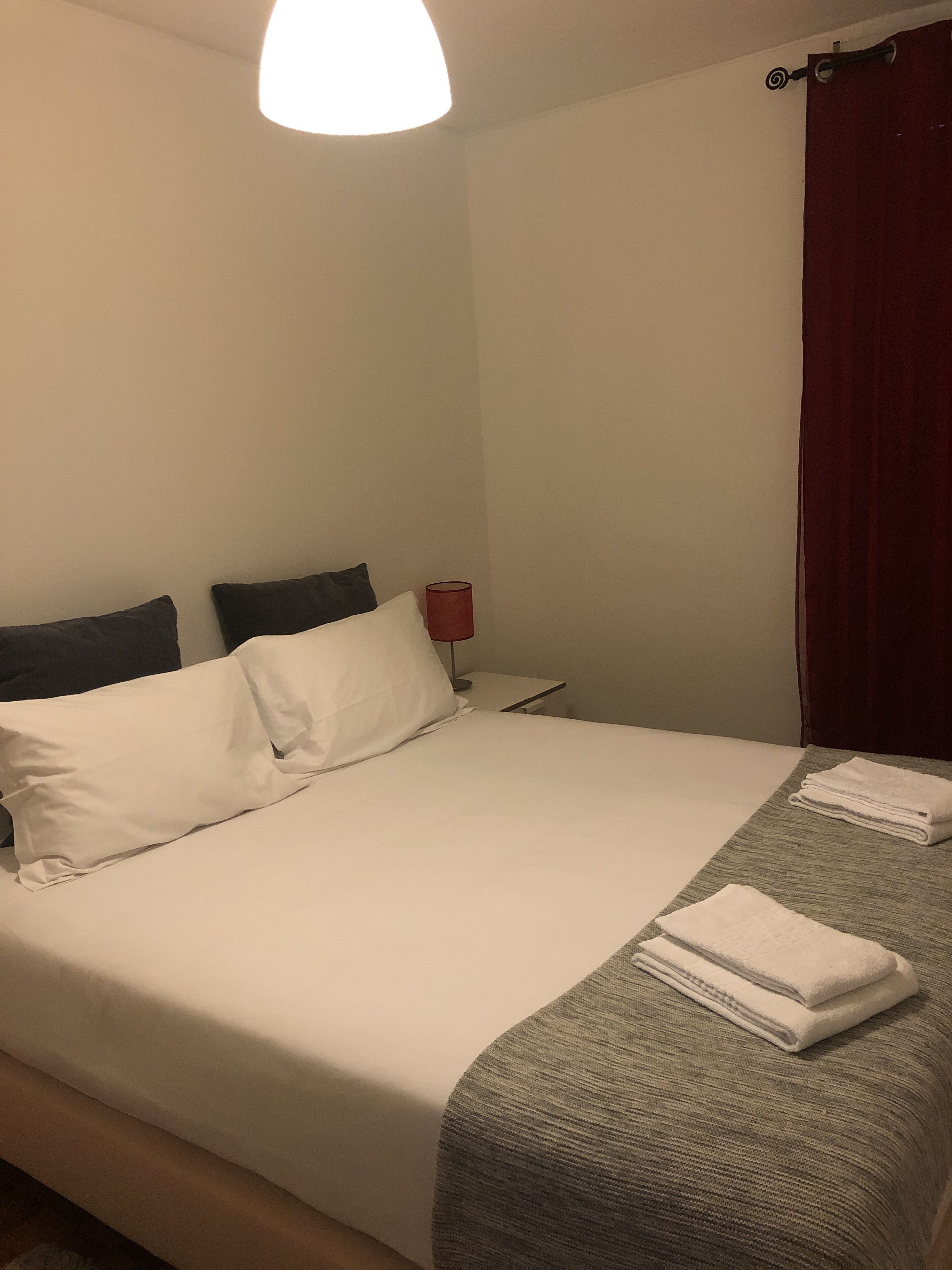 Un lit bien fait, avec du linge de lit blanc, un plaid gris à texture et deux oreillers. Une lampe rouge est posée sur une table de chevet, et un rideau rouge est partiellement visible.
