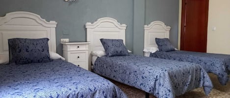Huoneessa on kolme sänkyä, joissa kaikissa on valkoinen pääty ja sinivalkoinen kuviollinen vuodevaatesetti. Huoneessa on myös puinen ovi ja pieni valkoinen yöpöytä, jossa on lamppu.