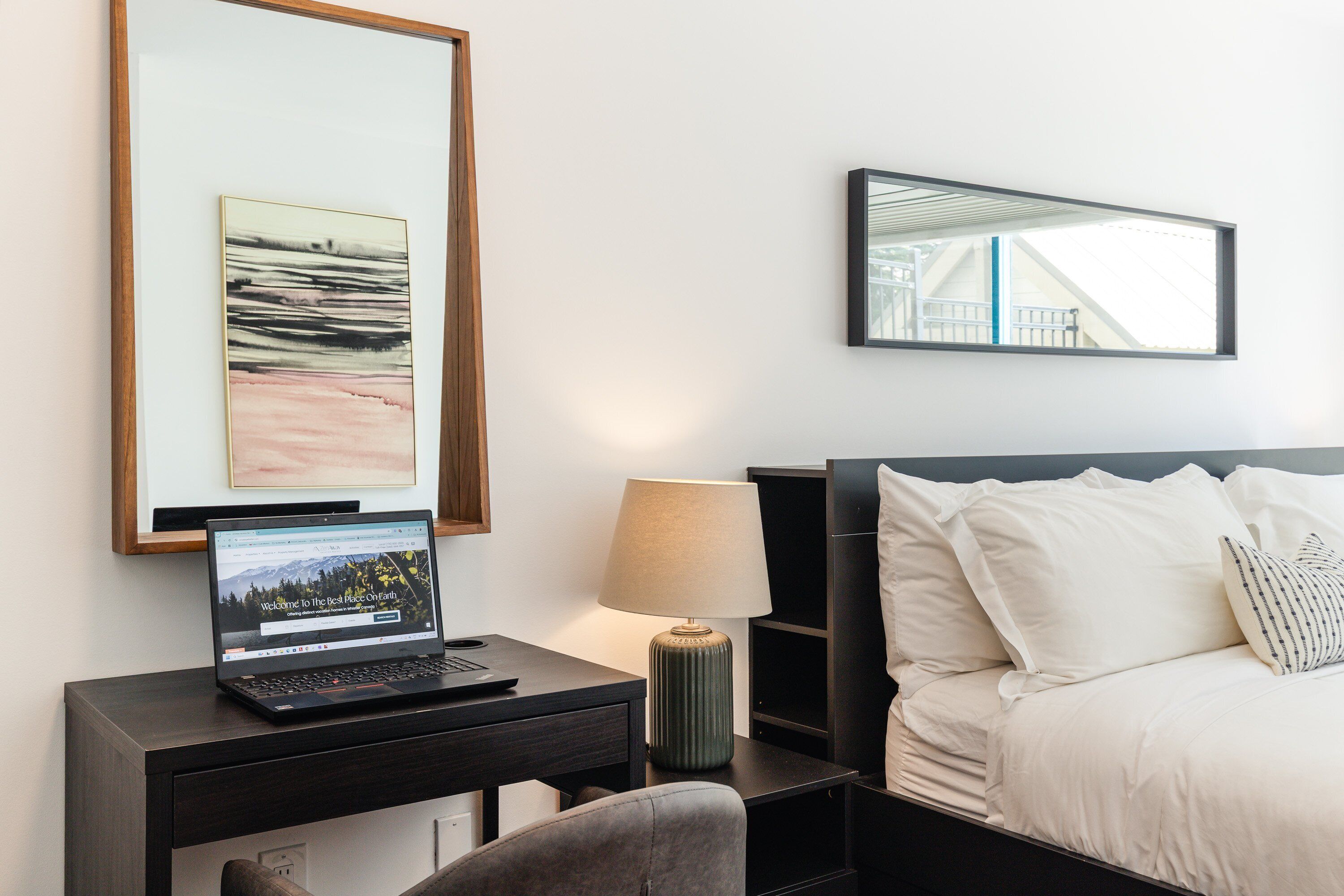 A hotel room with a bed, a desk with a laptop, a lamp, and two artworks on the wall.