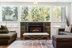 A living room with a fireplace, a sofa, a coffee table, and large windows with a forest view. - ❄︎Flute Escape❄︎ | 2 BR Ski In/Ski Out | Heated Pool & Hot Tub | Free Parking (Whistler)