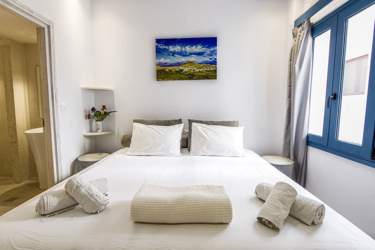 A hotel room with a bed, two pillows, a bedside table, a nightstand, a wall-mounted painting, a window with curtains, and a bathroom door.