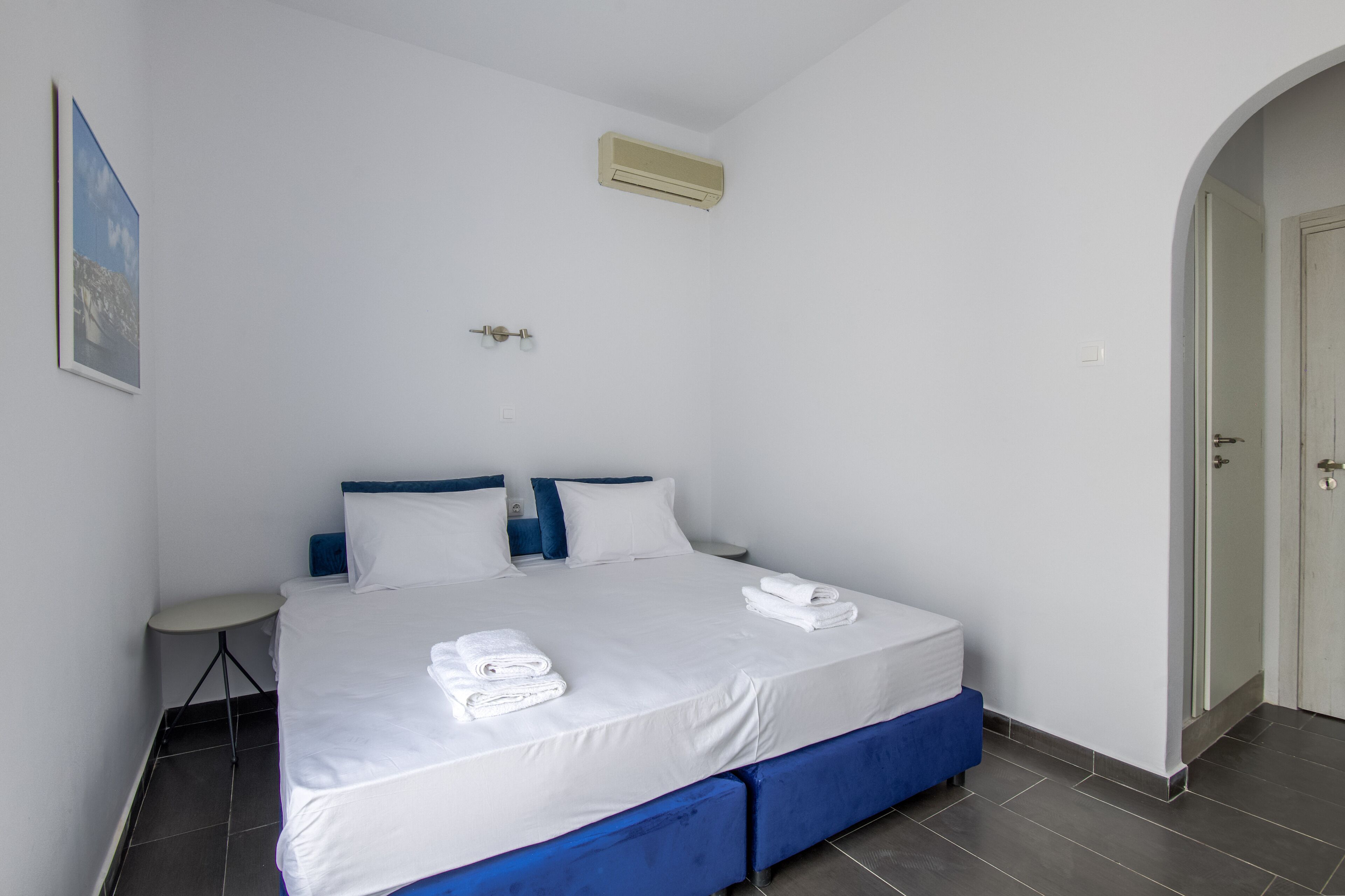A neatly made bed with white linens and blue headboard, a small bedside table, and a door to the right.