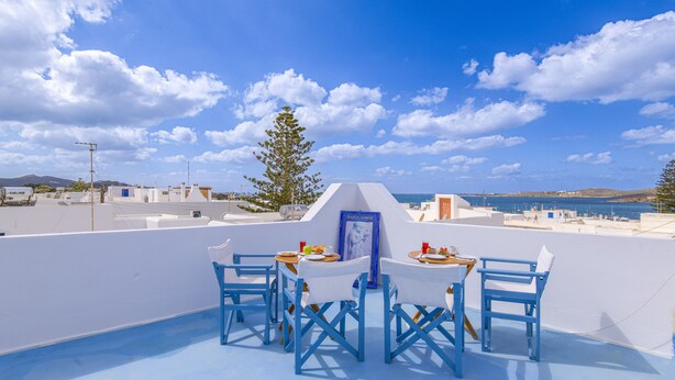 A rooftop terrace with blue chairs and tables, a framed picture, and a view of the sea and buildings. - Porto Bello Hotel (Paros)