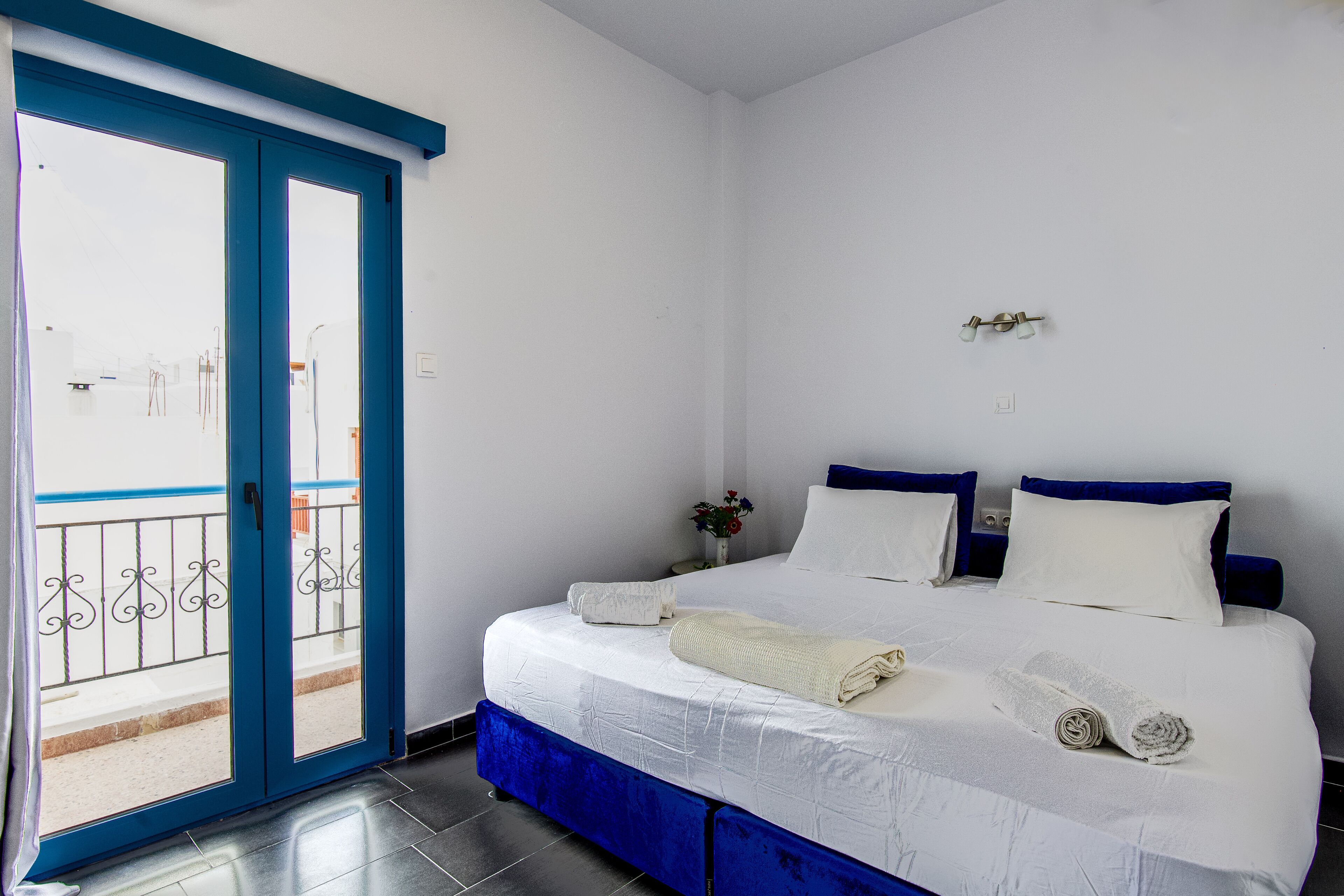 A bedroom with a bed, two pillows, a bedside table with a vase, and a blue-framed door leading to an outdoor area.