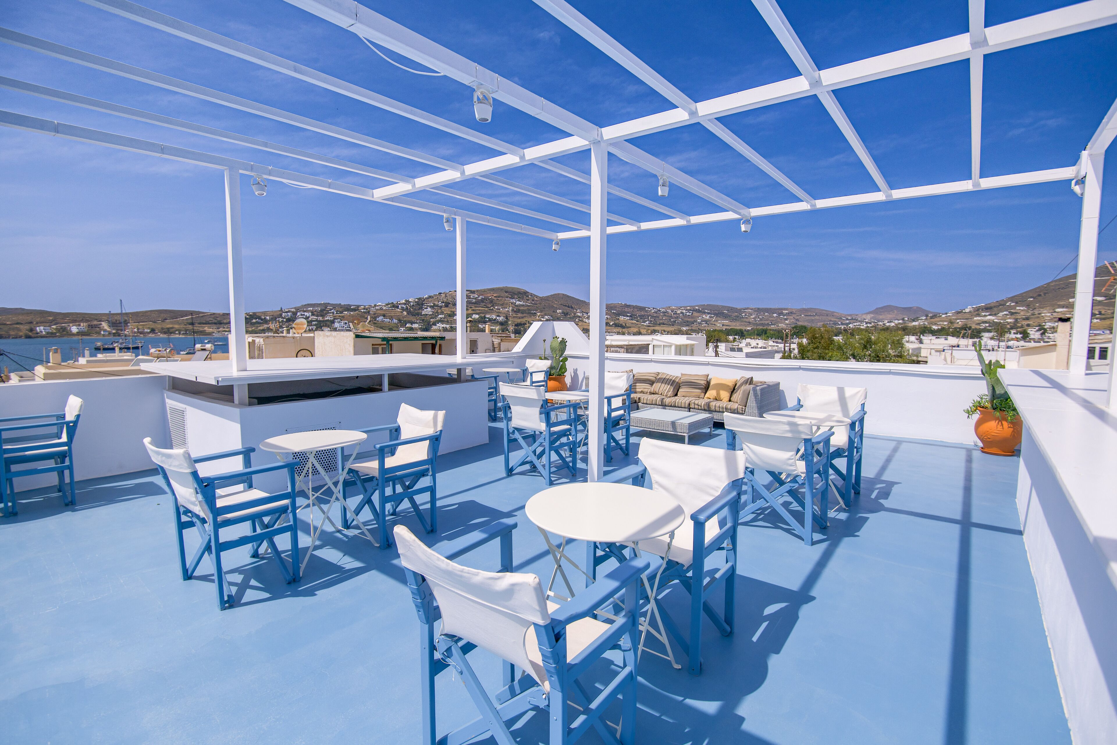 Una terraza en la azotea con mobiliario azul y blanco, una zona de bar y vistas a la costa y las colinas.