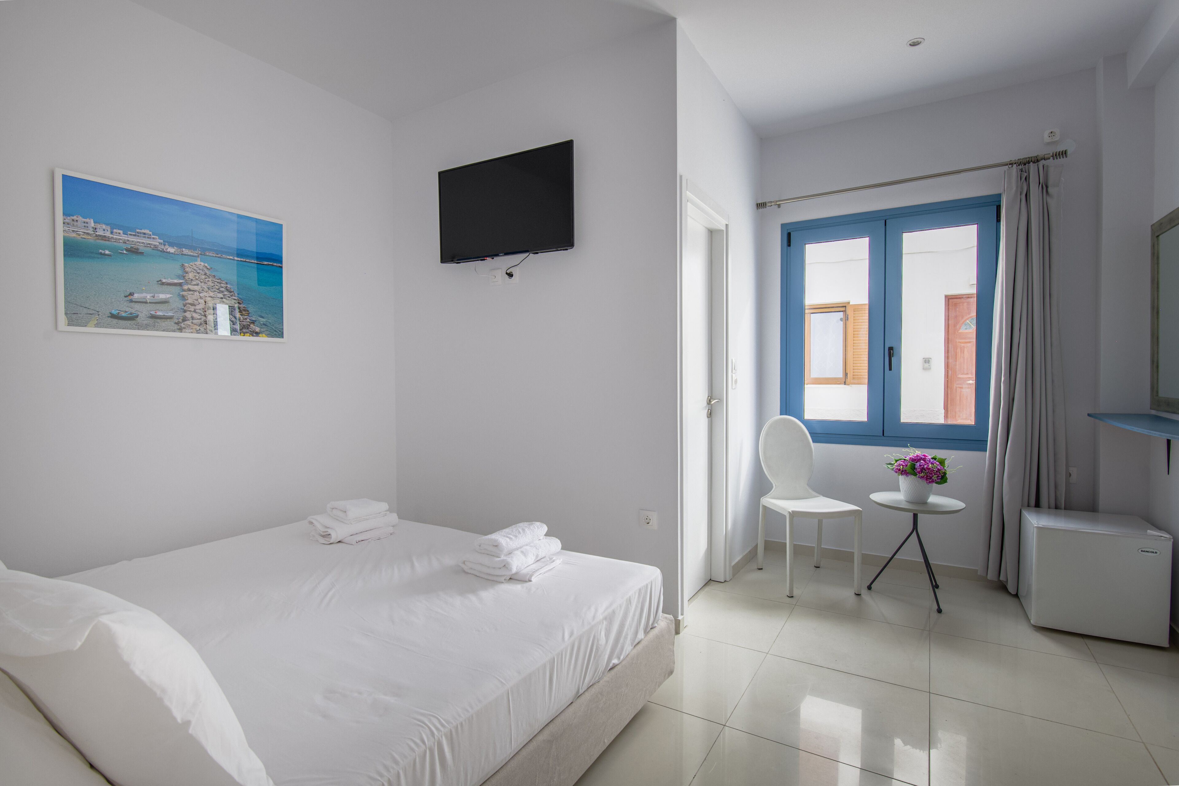 A hotel room with a bed, a TV mounted on the wall, a small table with a chair, and a window with blue shutters.