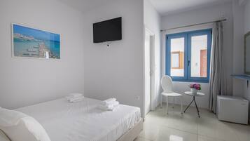 A hotel room with a bed, a TV mounted on the wall, a small table with a chair, and a window with blue shutters.