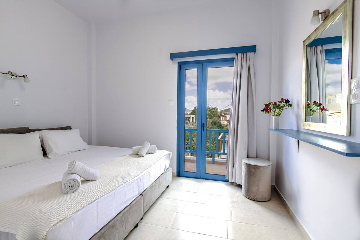 A bedroom with a bed, a small trash bin, a window with curtains, and a mirror on the wall.