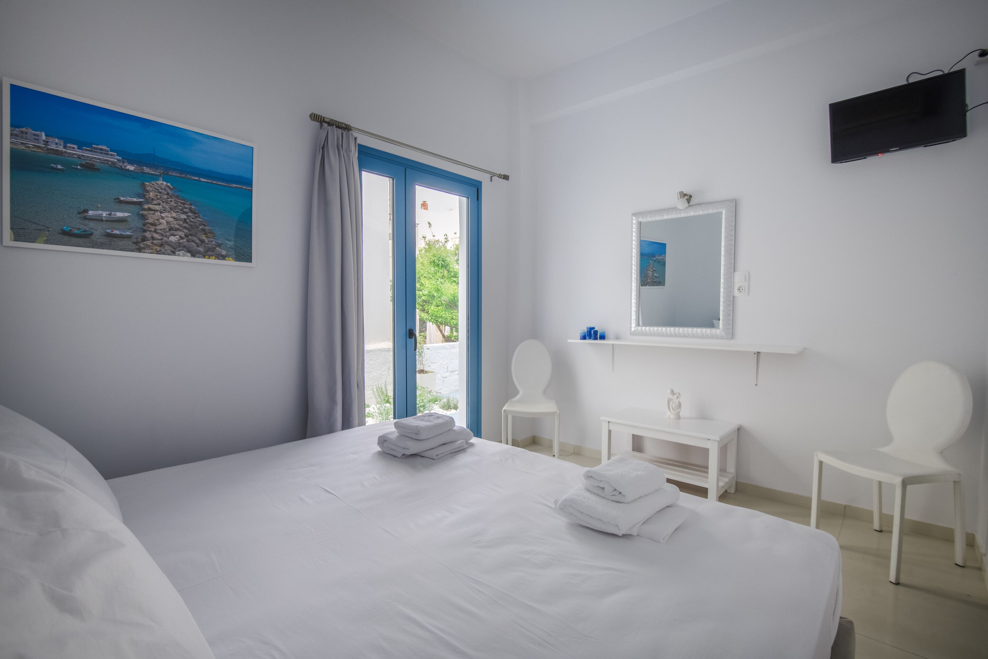 Ein Hotelzimmer mit einem Bett, einem Schreibtisch, zwei Stühlen, einem Fernseher, einem Gemälde und Meerblick durch ein Fenster.