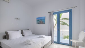 A bedroom with a bed, a chair, a window with curtains, a picture on the wall, and a view of the beach. - Porto Bello Hotel (Paros)