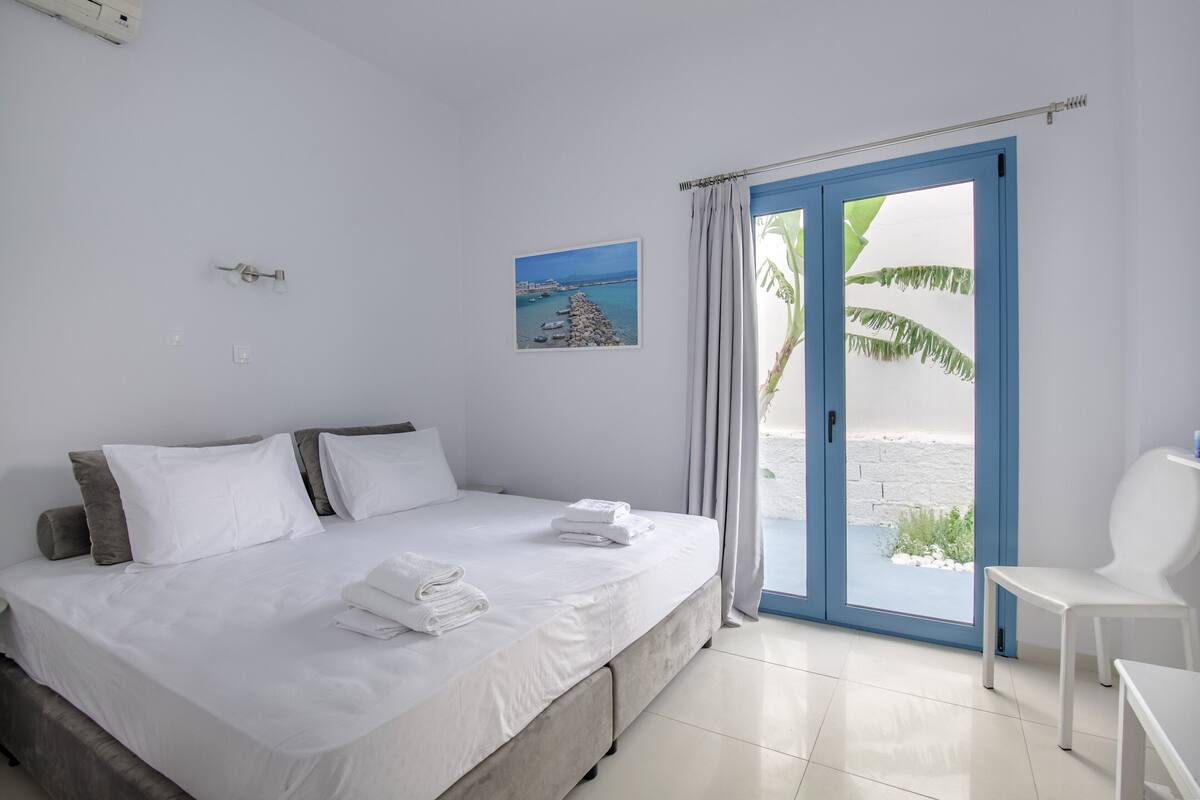 A bedroom with a bed, a chair, a window with curtains, a picture on the wall, and a view of the beach.