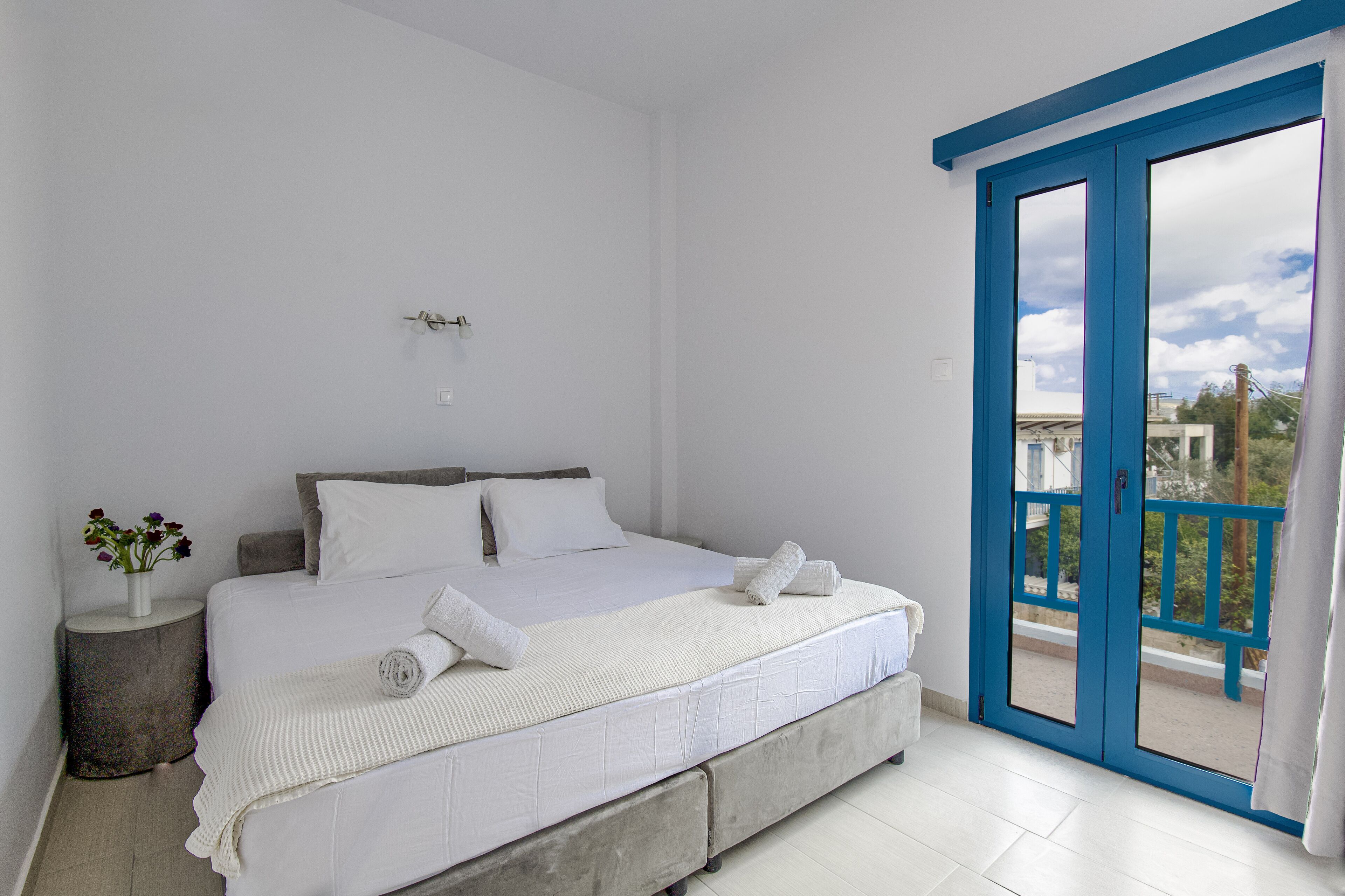 Un dormitorio con una cama, una mesita de noche con un florero y un balcón con puertas azules.