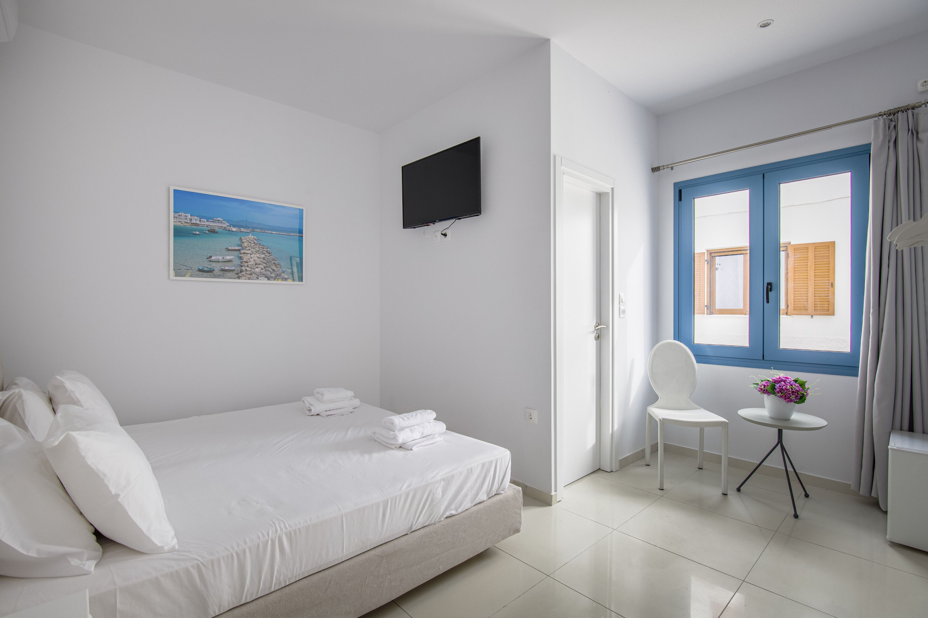 Ein Hotelzimmer mit Bett, einem an der Wand befestigten Fernseher, einem Gemälde einer Küstenlandschaft, einem Fenster mit blauen Fensterläden und einem kleinen Tisch mit Blumenvase.