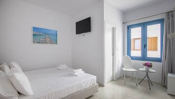 Ein Hotelzimmer mit Bett, einem an der Wand befestigten Fernseher, einem Gemälde einer Küstenlandschaft, einem Fenster mit blauen Fensterläden und einem kleinen Tisch mit Blumenvase.