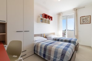 Quarto com duas camas, uma escrivaninha e uma cadeira. Há uma janela com cortinas e um quadro na parede.