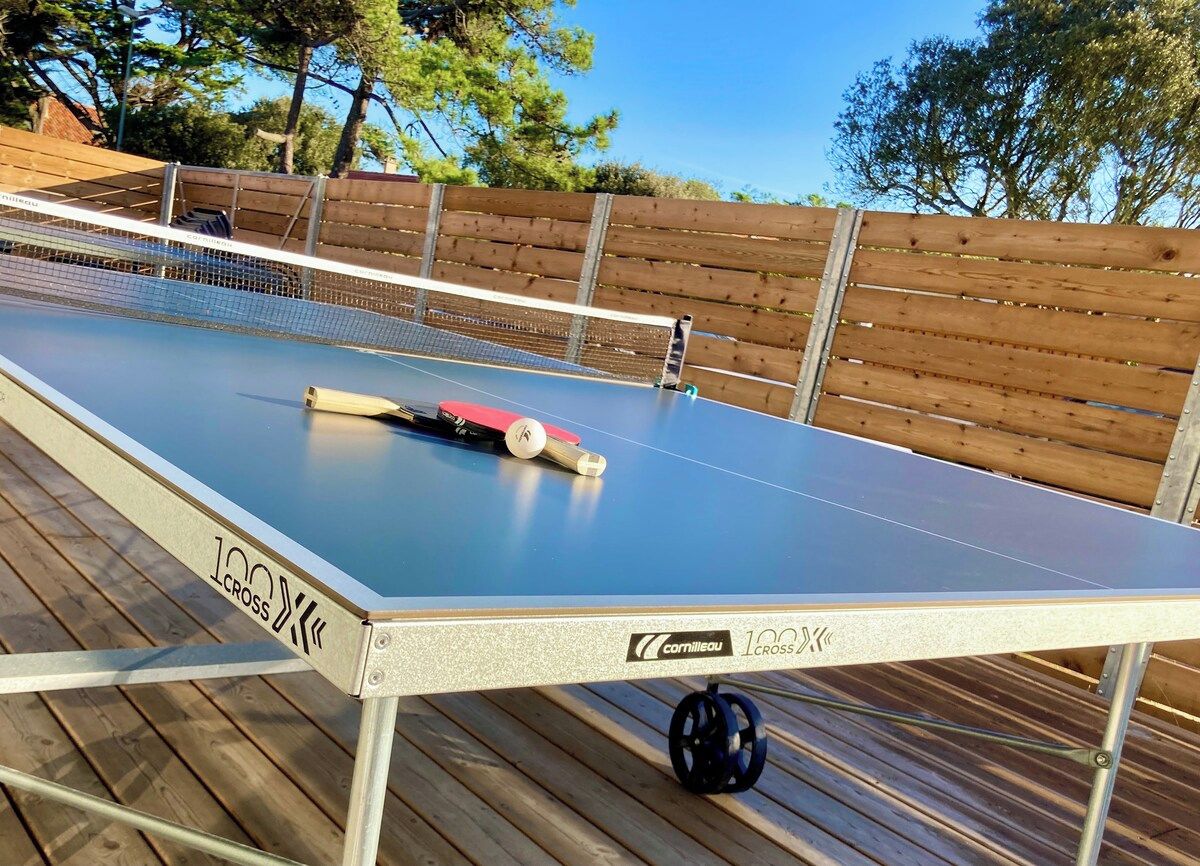 Et bordtennisbord med net, placeret udendørs på en træterrasse med et træhegn i baggrunden.