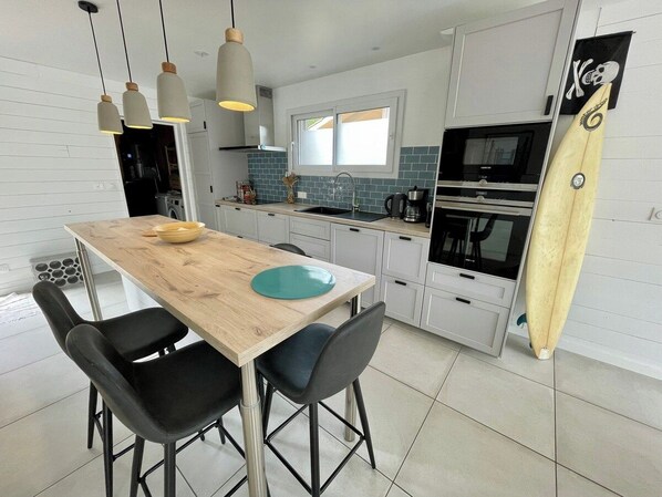 Una cocina moderna con una mesa de madera, sillas negras y una tabla de surf apoyada en la pared.