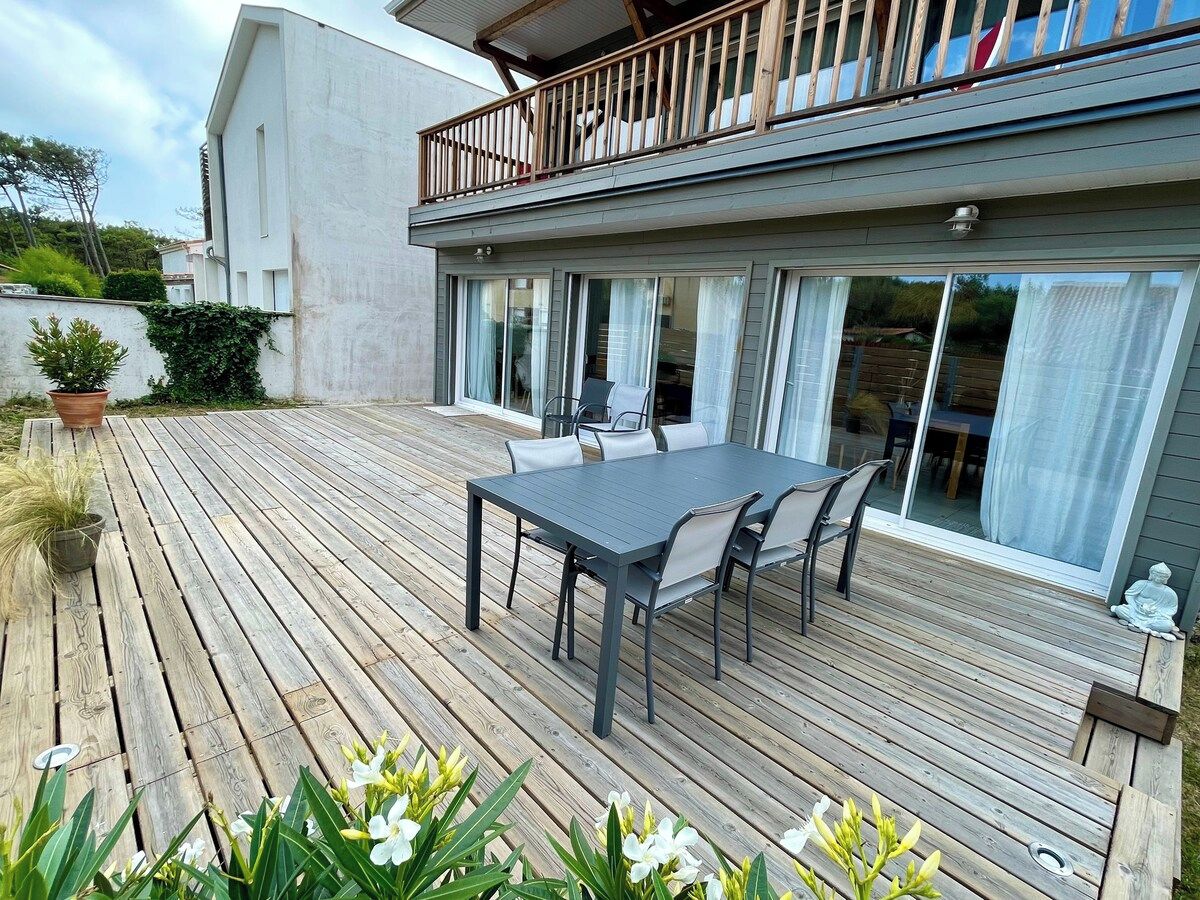 A wooden deck with a table and chairs, a balcony with a railing, and a building with large windows.