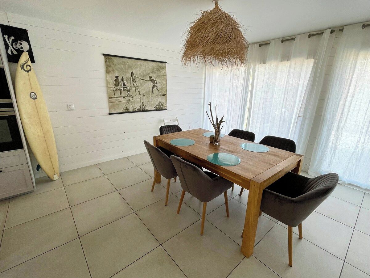 A dining area with a wooden table, six chairs, and a surfboard leaning against the wall.