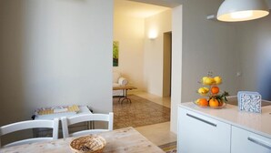 Una cocina moderna con armarios blancos, una mesa de comedor con sillas, una lámpara colgante y una exposición de frutas en la encimera.