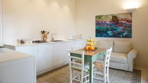 Una cocina con armarios blancos, una mesa de comedor con sillas, un cuadro en la pared y piso de baldosas.