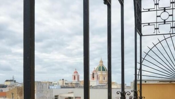 Un balcón con barandillas de metal decorativo y vista a edificios en el fondo.