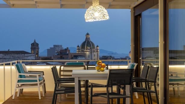 A dining area with a table and chairs, a decorative hanging light, and a view of a cityscape with buildings and a dome structure. - Terrammare apartment (Trapani)