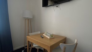 Una habitación pequeña con una mesa de madera, una única silla blanca, una lámpara de pie y un televisor de pantalla plana montado en la pared.