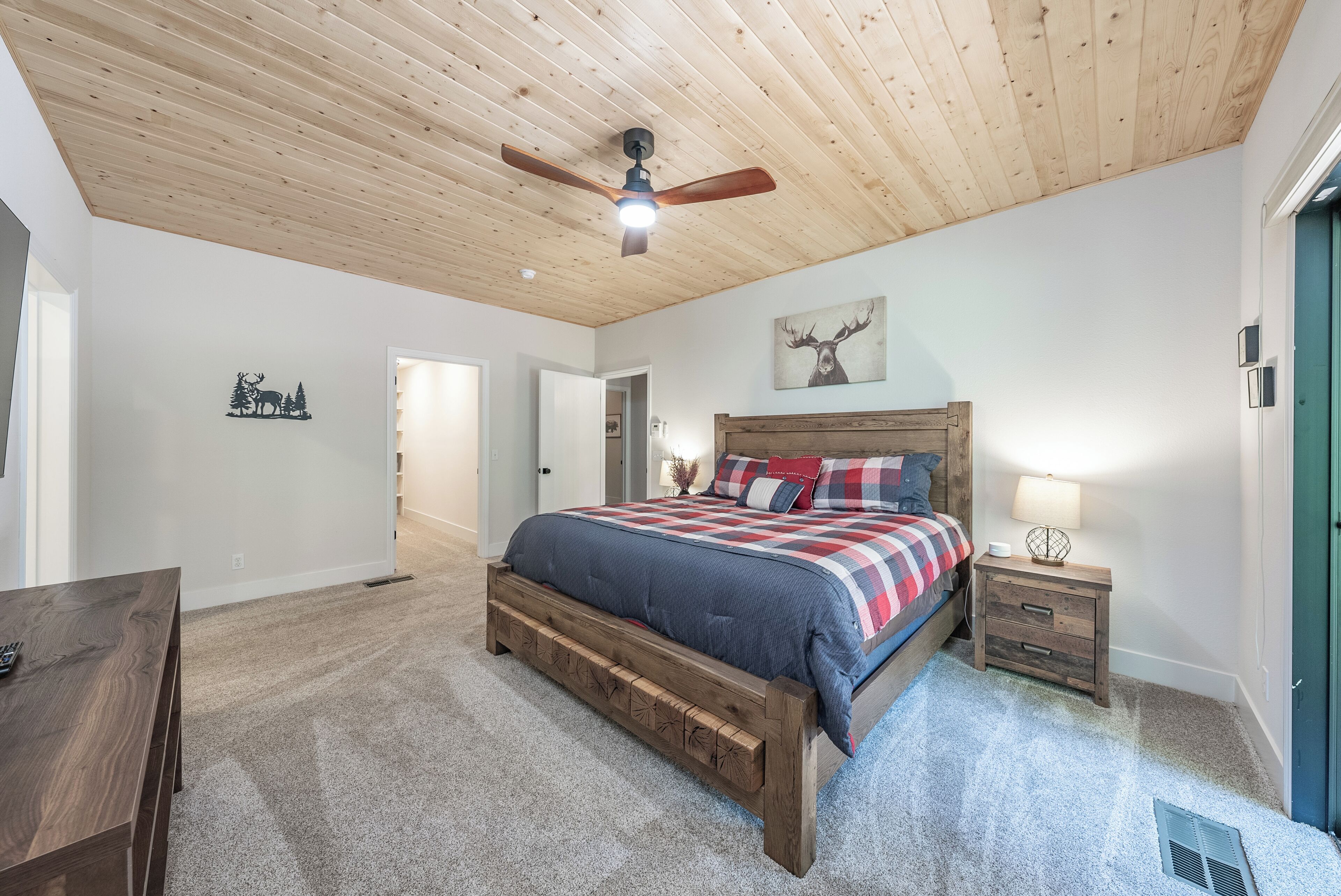 A bedroom with a wooden ceiling, a bed with a plaid bedspread, a nightstand with a lamp, and a wall-mounted deer artwork.