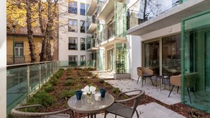 Un patio exterior moderno con una mesa redonda y sillas de mimbre, rodeado por barandas de vidrio y un jardín con plantas y hojas secas.