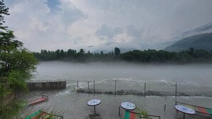 Uitzicht op een rivier met een houten terras en tafels, omgeven door groen en bergen.