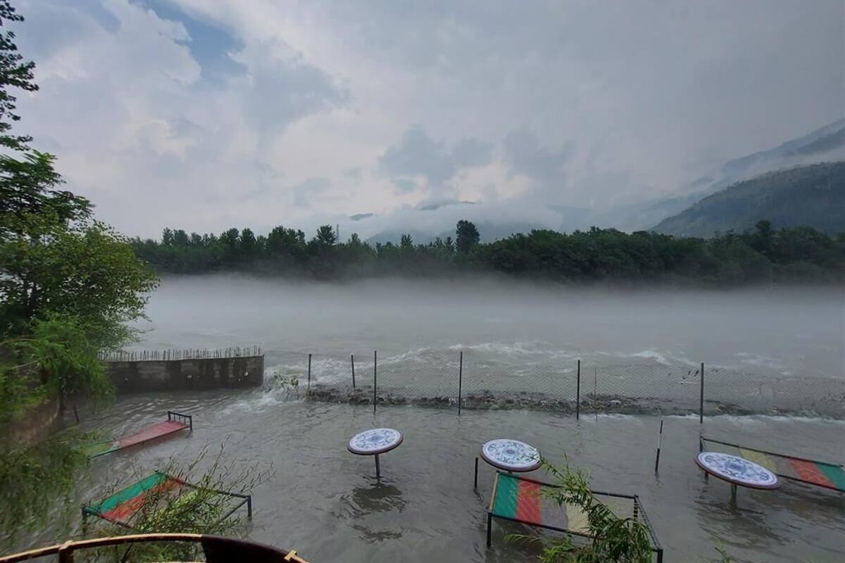 Uitzicht op een rivier met een houten terras en tafels, omgeven door groen en bergen.