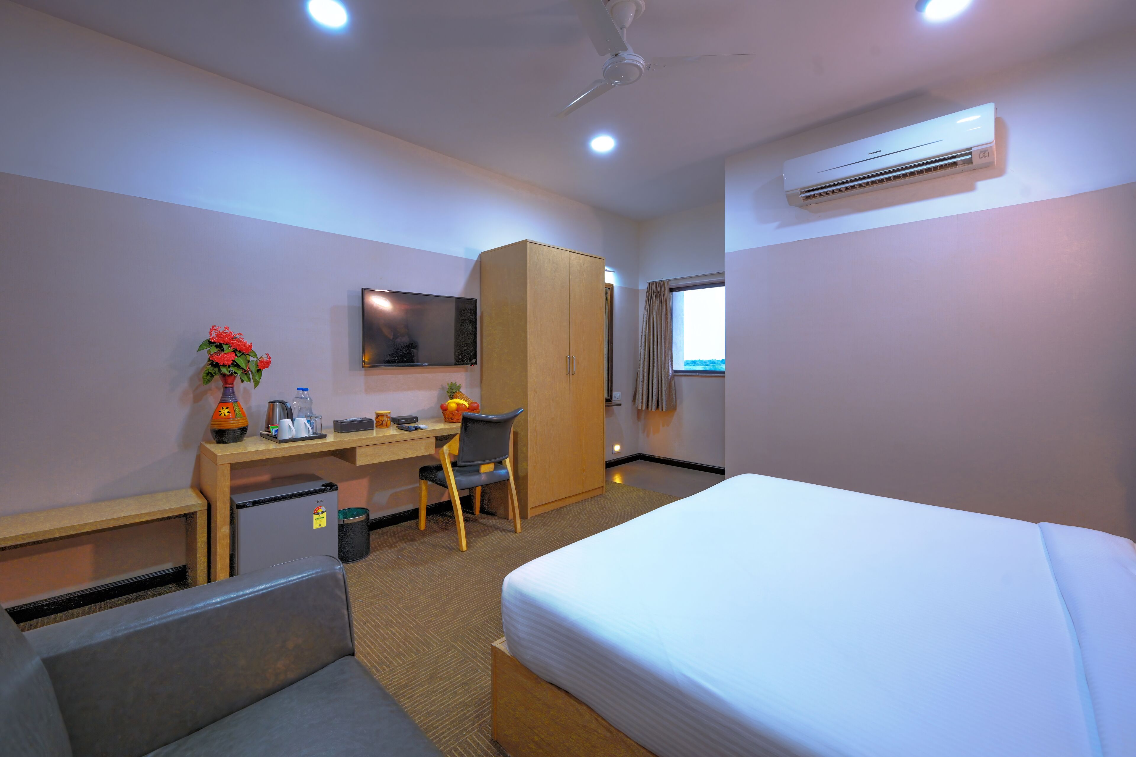 A hotel room with a bed, a desk with a chair, a television, an air conditioner, and a wardrobe.