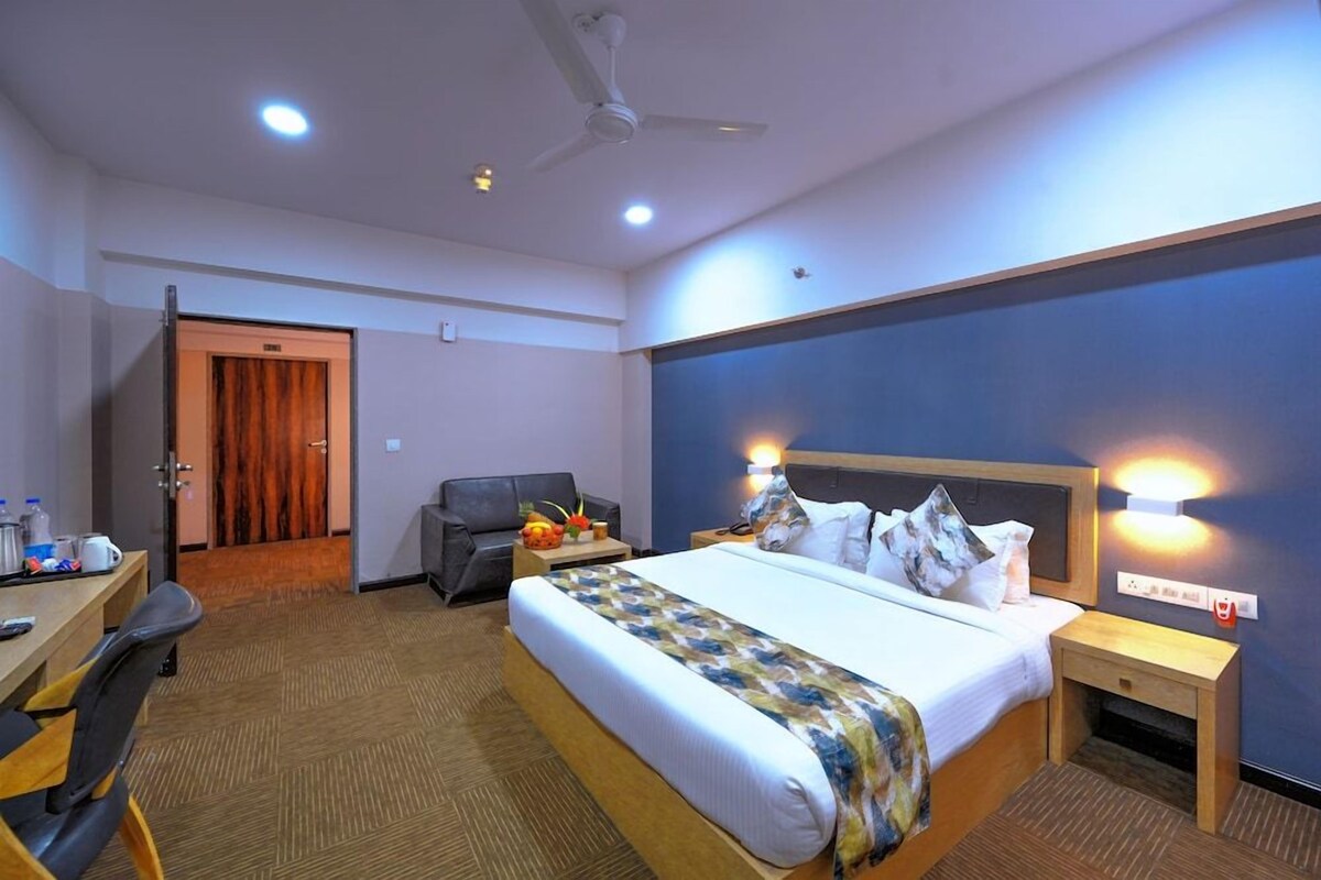 A hotel room with a bed, a desk, a chair, a ceiling fan, and a door leading to another room.