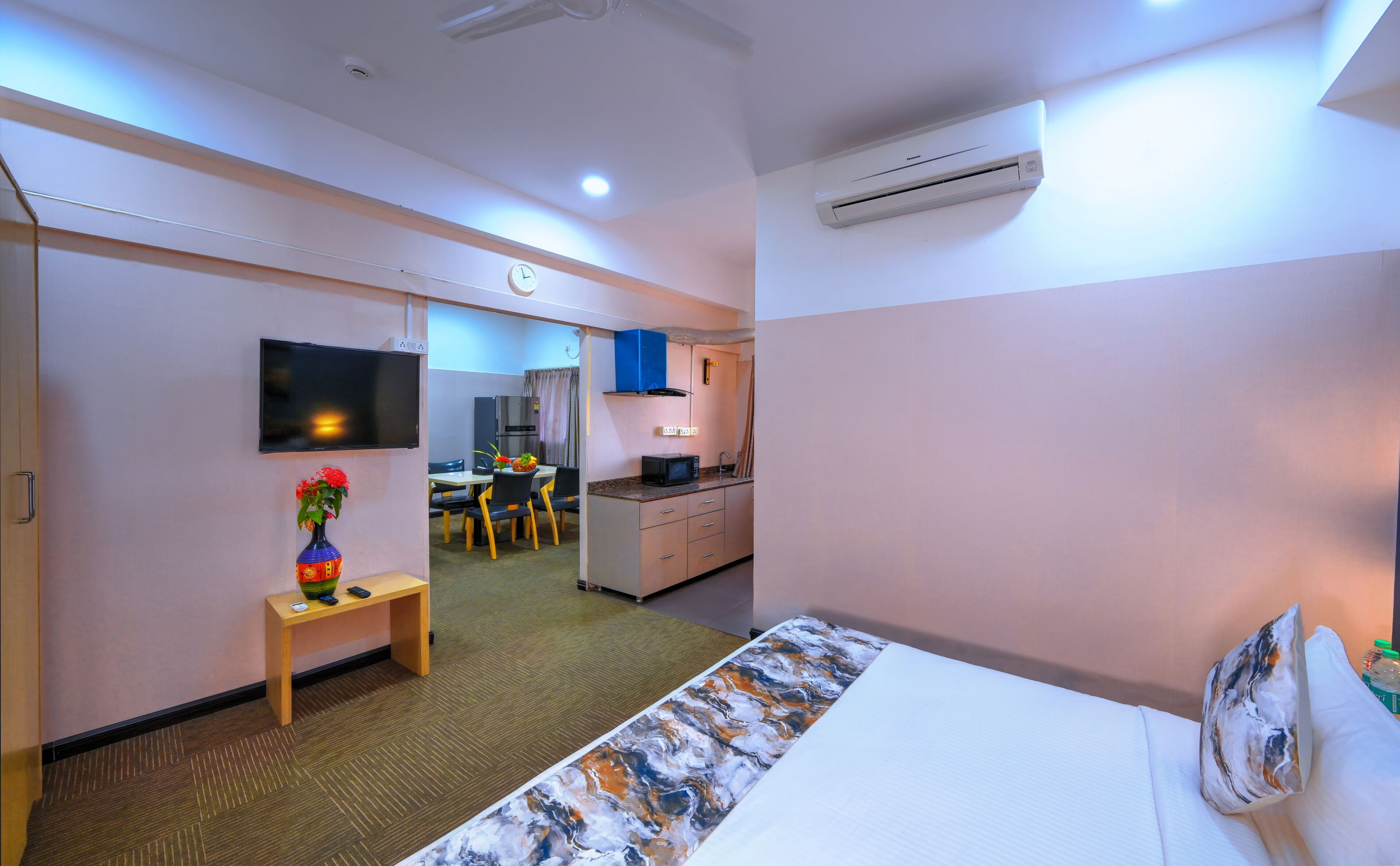 Habitación de hotel con cama, escritorio con un florero, televisor, kitchenette y baño visible al fondo.