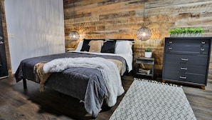 A bedroom with a wooden wall, a bed with a grey bedspread, two bedside lamps, a small nightstand with a plant, and a dark dresser. - Best of the Smokies! •Game Room•GSMNP•Theater•Sleep 16•Hot Tub•Flat Parking (Sevierville)