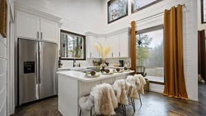 A modern kitchen with white cabinetry, a stainless steel refrigerator, a central island with bar stools, and a view through large windows. - Moroccan Nights|Hot Tub|Fire Pit|Hammocks (Broken Bow)