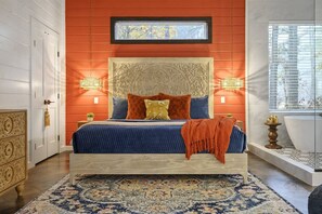 A bedroom with a large bed, a headboard with intricate carvings, two bedside lamps, a window with blinds, and a bathroom with a bathtub.