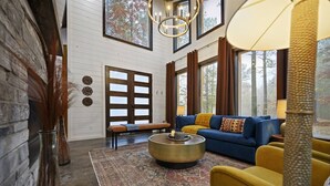 A cozy living room with a blue sofa, yellow armchair, and a round coffee table. There is a large window with curtains, a fireplace, and a rug on the floor. - Moroccan Nights|Hot Tub|Fire Pit|Hammocks (Broken Bow)